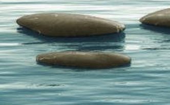 Das Bild zeigt drei große, flache Steine, die aus einer ruhigen Wasseroberfläche herausragen. Die Steine sind dunkelgrau und haben eine ovale Form. Das Wasser hat eine blaue Farbe mit sanften Wellen, die sich um die Steine kräuseln. Die Farbe des Wassers wird nach hinten hin heller, was für eine ruhige und friedliche Atmosphäre sorgt.